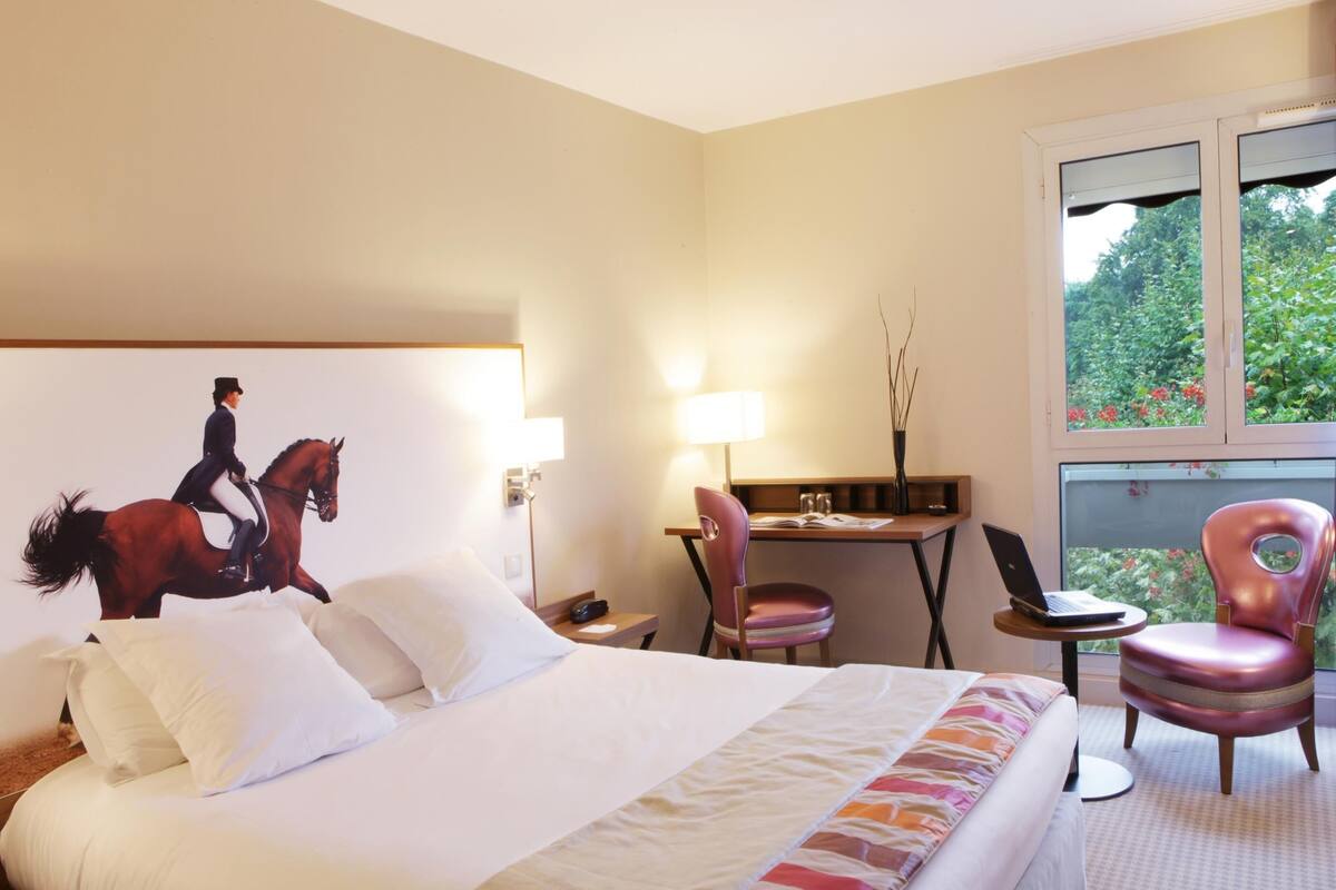 A hotel room with a bed, a desk, a chair, and a window with a view of greenery.