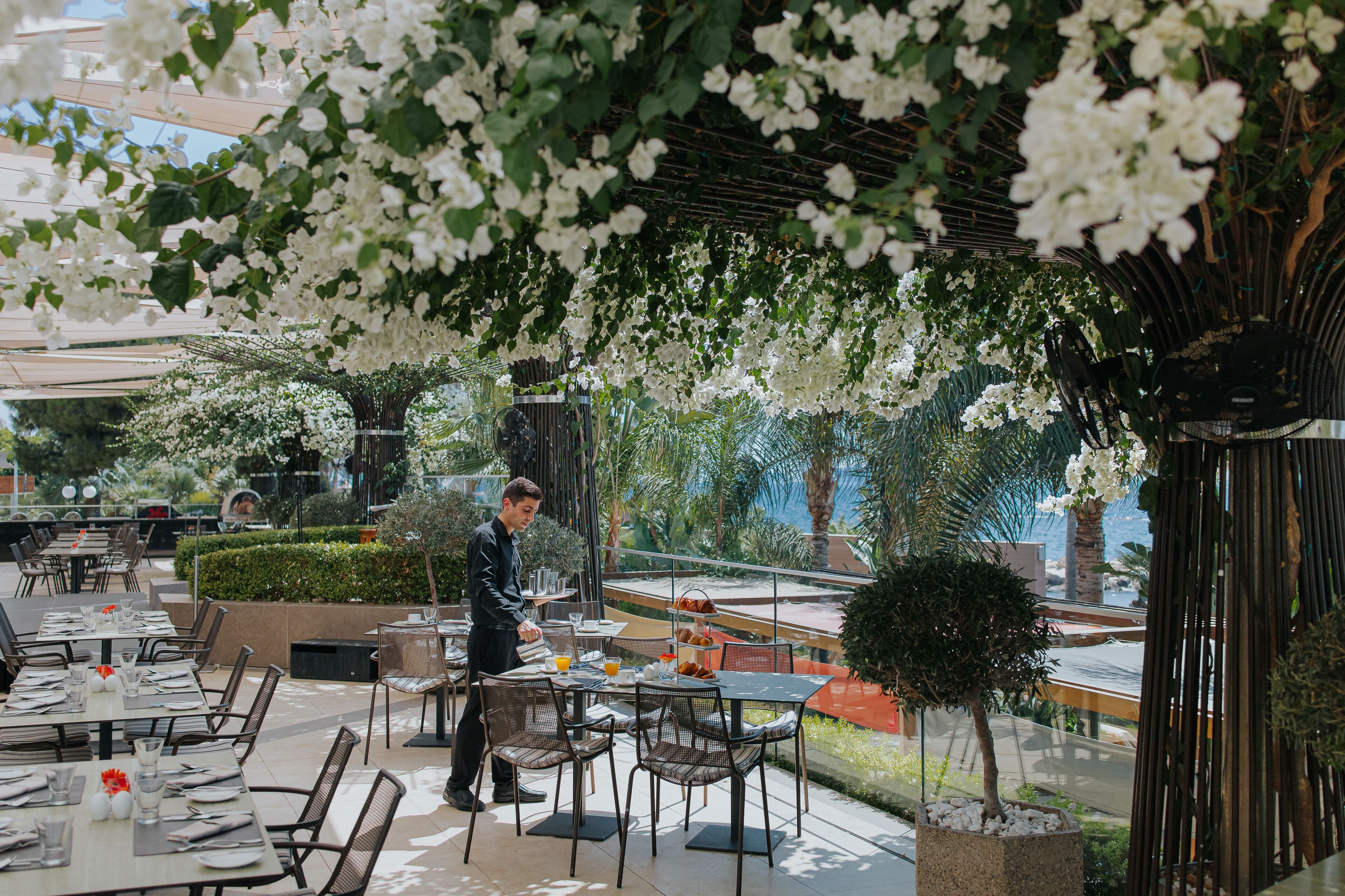 An outdoor dining area with tables and chairs, a waiter, and a view of the sea.
