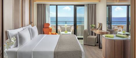 A hotel room with a large bed, a desk with a chair, a small table with a chair, and a view of the ocean through large windows.