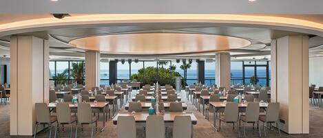 A large conference room with multiple tables and chairs, large windows with a view of the ocean, and a curved ceiling design.