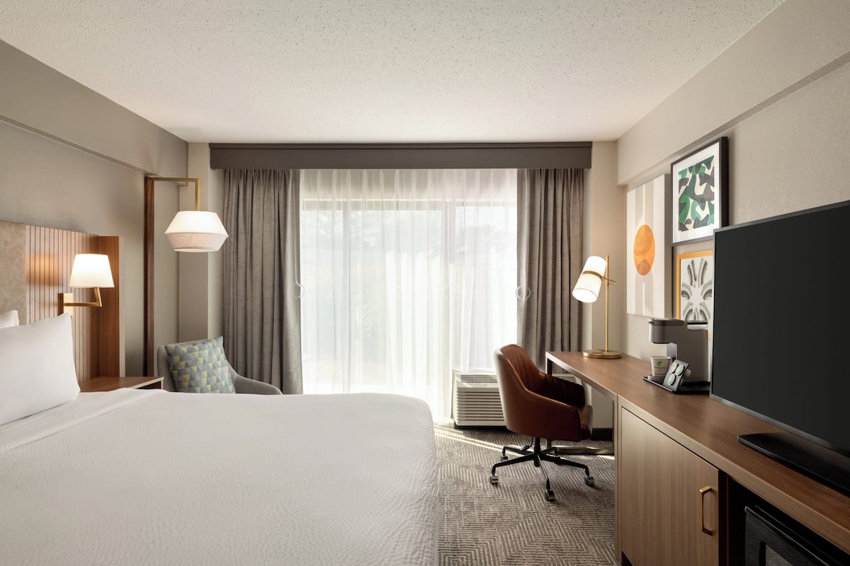 A hotel room with a large bed, a desk with a flat-screen TV, a chair, and a desk lamp.
