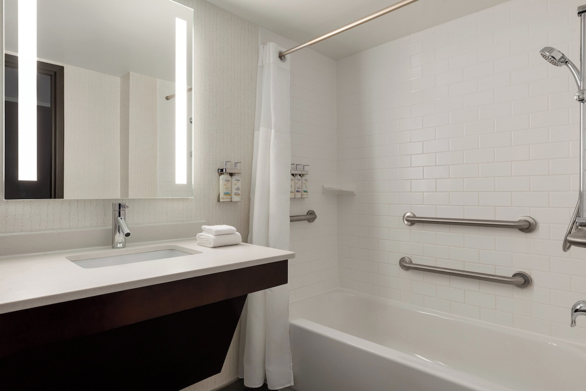 Ein modernes Badezimmer mit Waschbecken, Spiegel und Badewanne. Es sind Haltegriffe zur Sicherheit angebracht, und eine Duschbrause ist sichtbar.