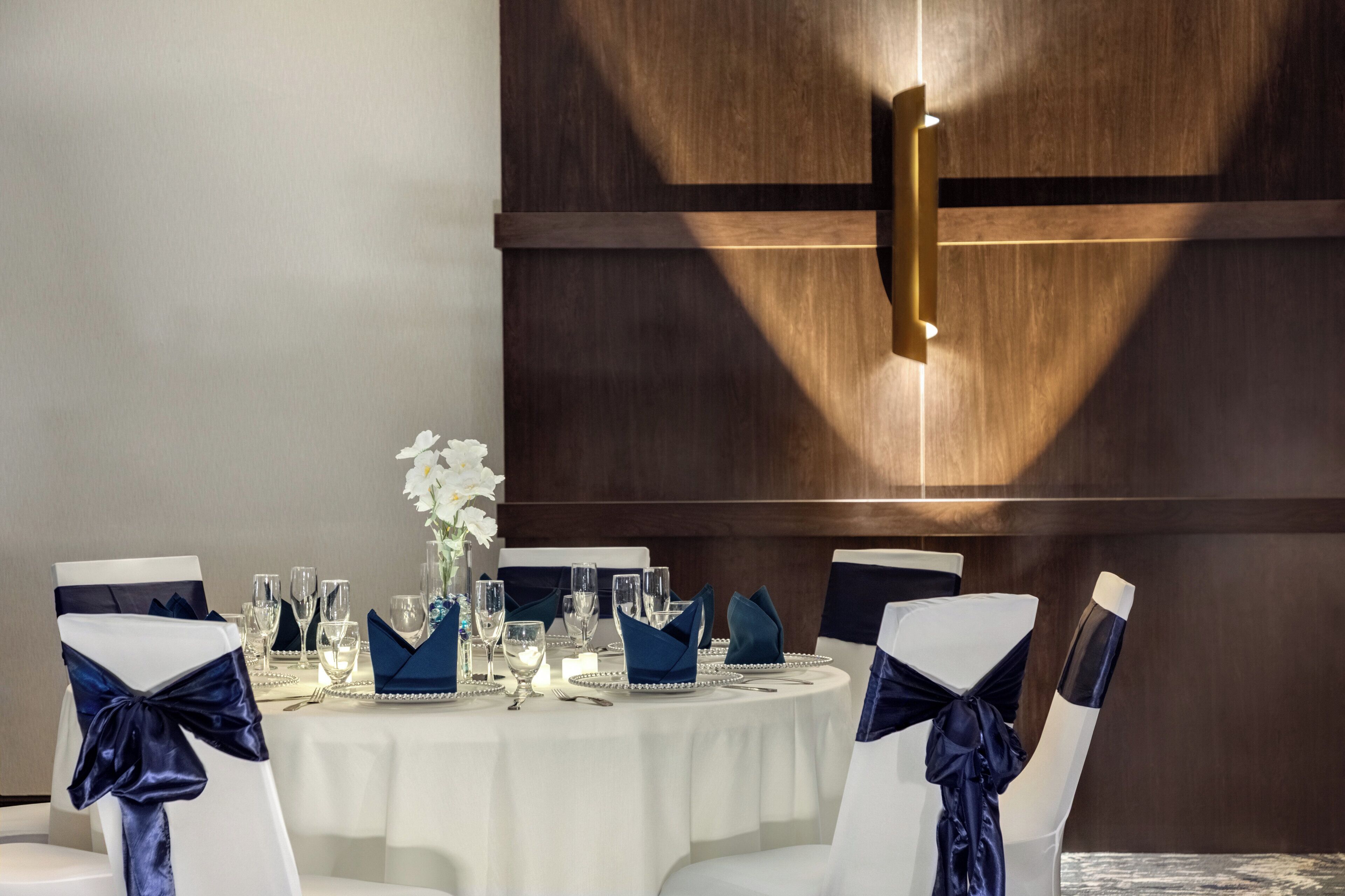 A round table with white chairs, each adorned with a dark blue ribbon. The table is set with glassware, a vase with white flowers, and a candle.