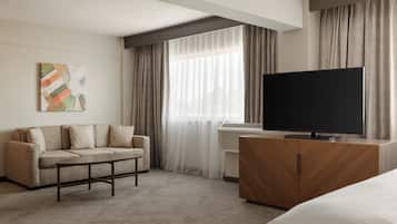 A hotel room with a sofa, a flat-screen TV, a wooden cabinet, and a large window with sheer curtains.