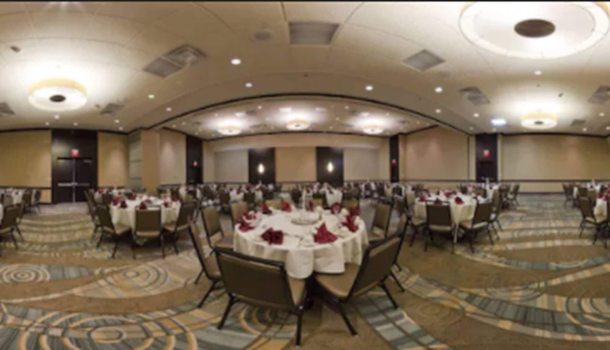 Un amplio salón de banquetes con mesas redondas dispuestas para comer, pisos alfombrados y un alto techo con iluminación empotrada.