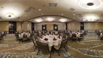 Un amplio salón de banquetes con mesas redondas dispuestas para comer, pisos alfombrados y un alto techo con iluminación empotrada.
