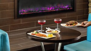Uma mesa de jantar com uma refeição e dois copos de bebida vermelha, uma lareira acesa e cadeiras azuis.