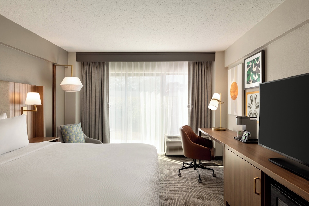 Ein Hotelzimmer mit einem großen Bett, einem Schreibtisch mit Flachbildfernseher, einem Sessel und einer Schreibtischlampe.