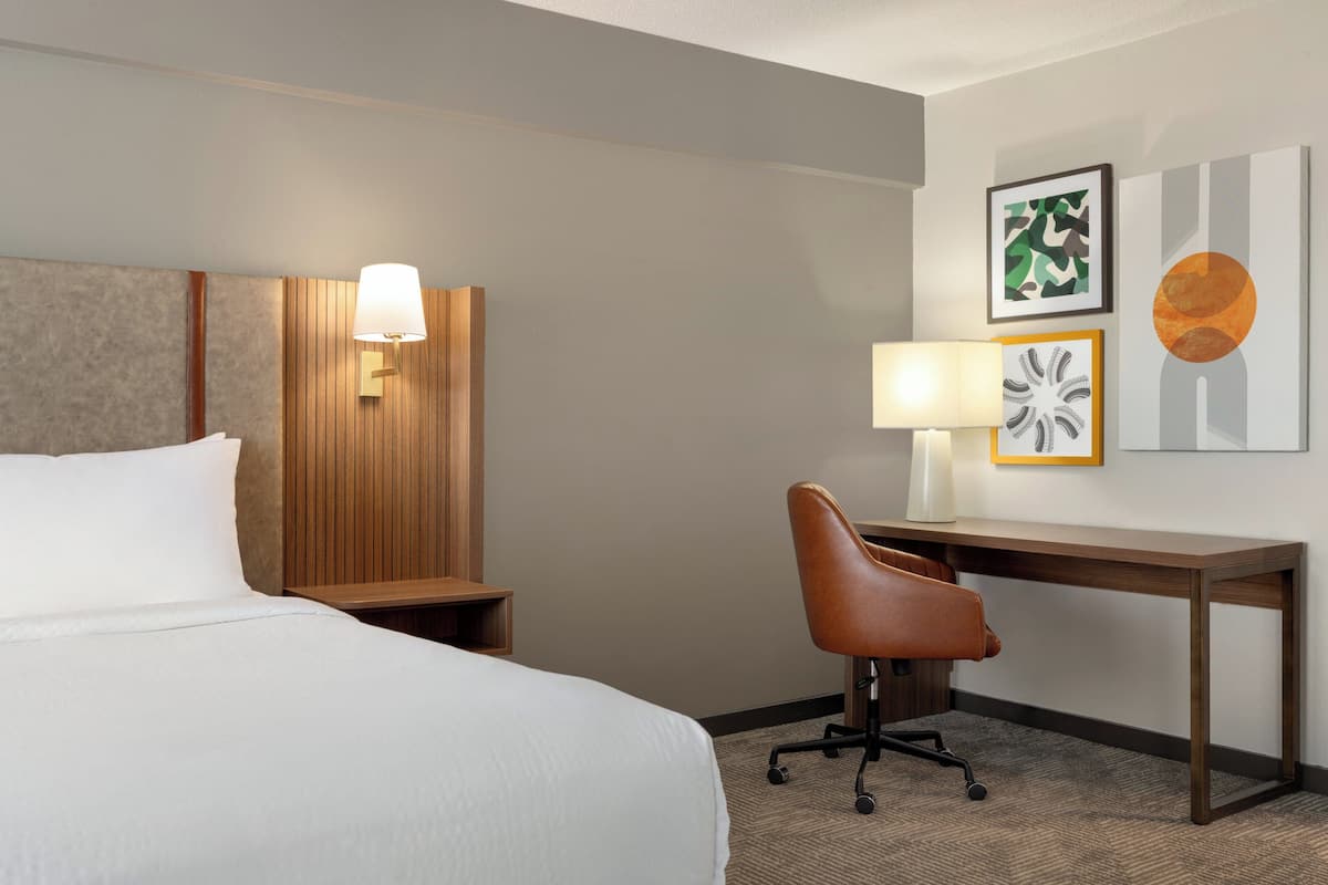 A hotel room with a bed, a desk, a chair, a lamp, and artwork on the wall.