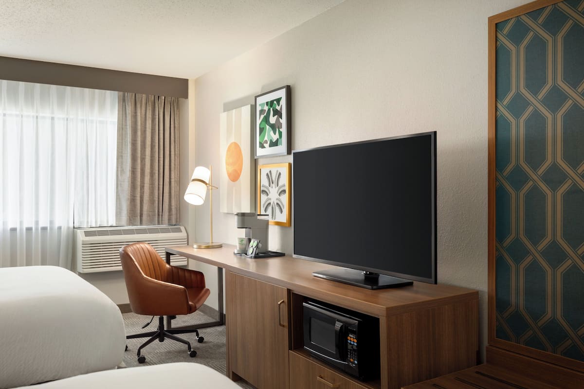 A hotel room with a bed, a desk with a flat-screen TV, a microwave, a chair, and a lamp.