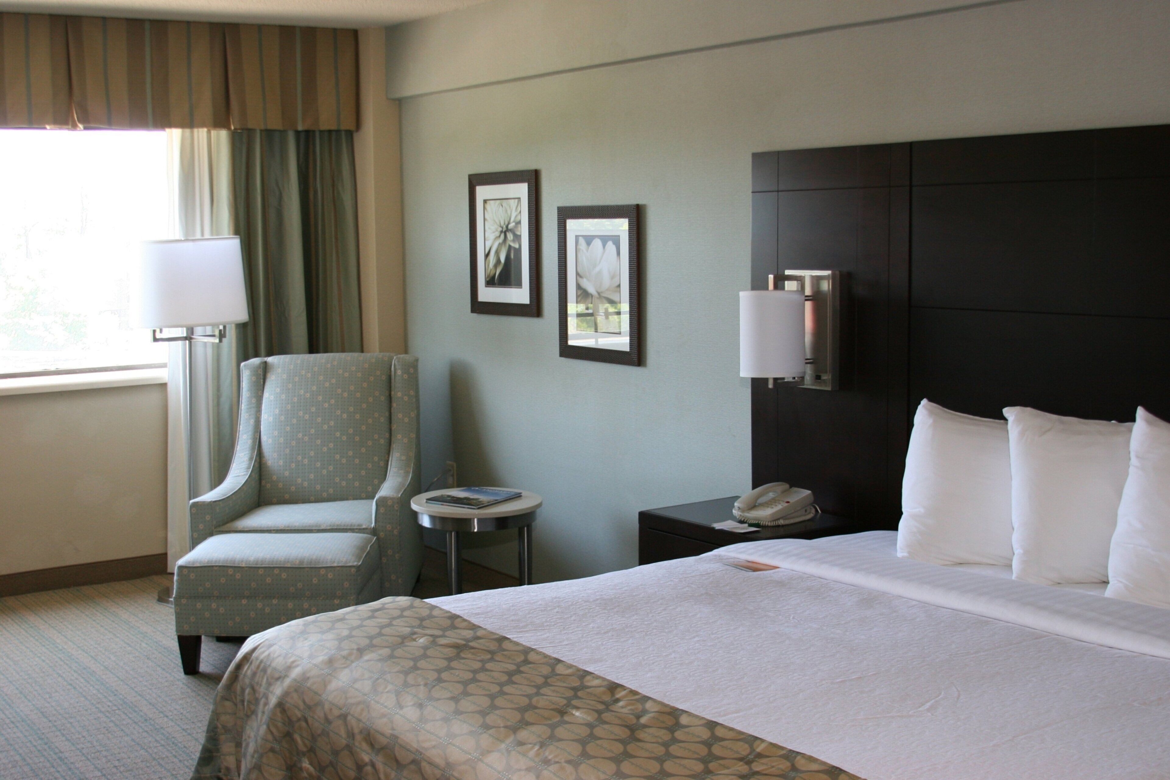 A hotel room with a bed, a chair, a bedside table, a lamp, and two framed pictures on the wall.