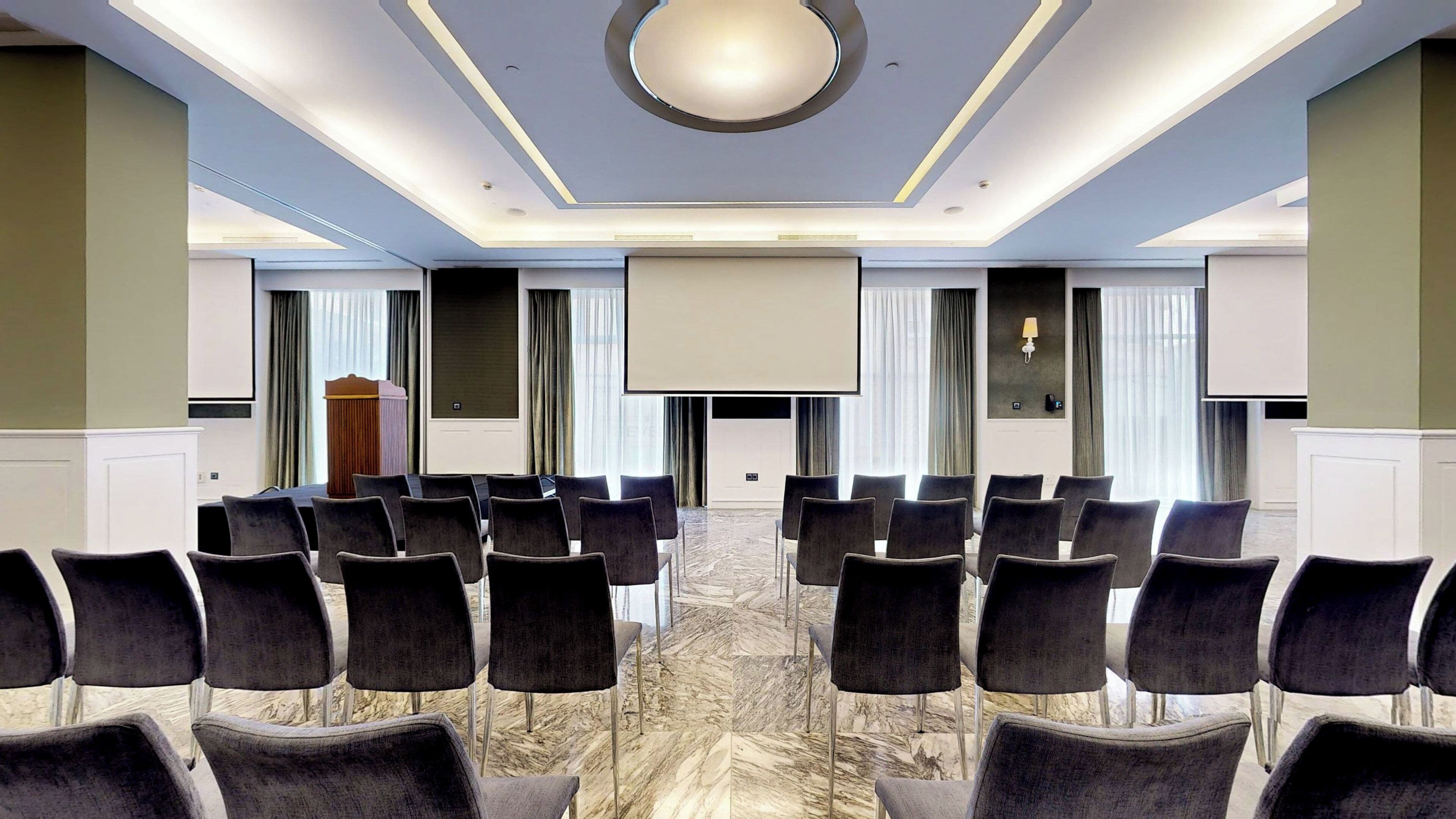 Un salón de conferencias con filas de sillas mirando hacia una pantalla grande, un atril y un piso de mármol.