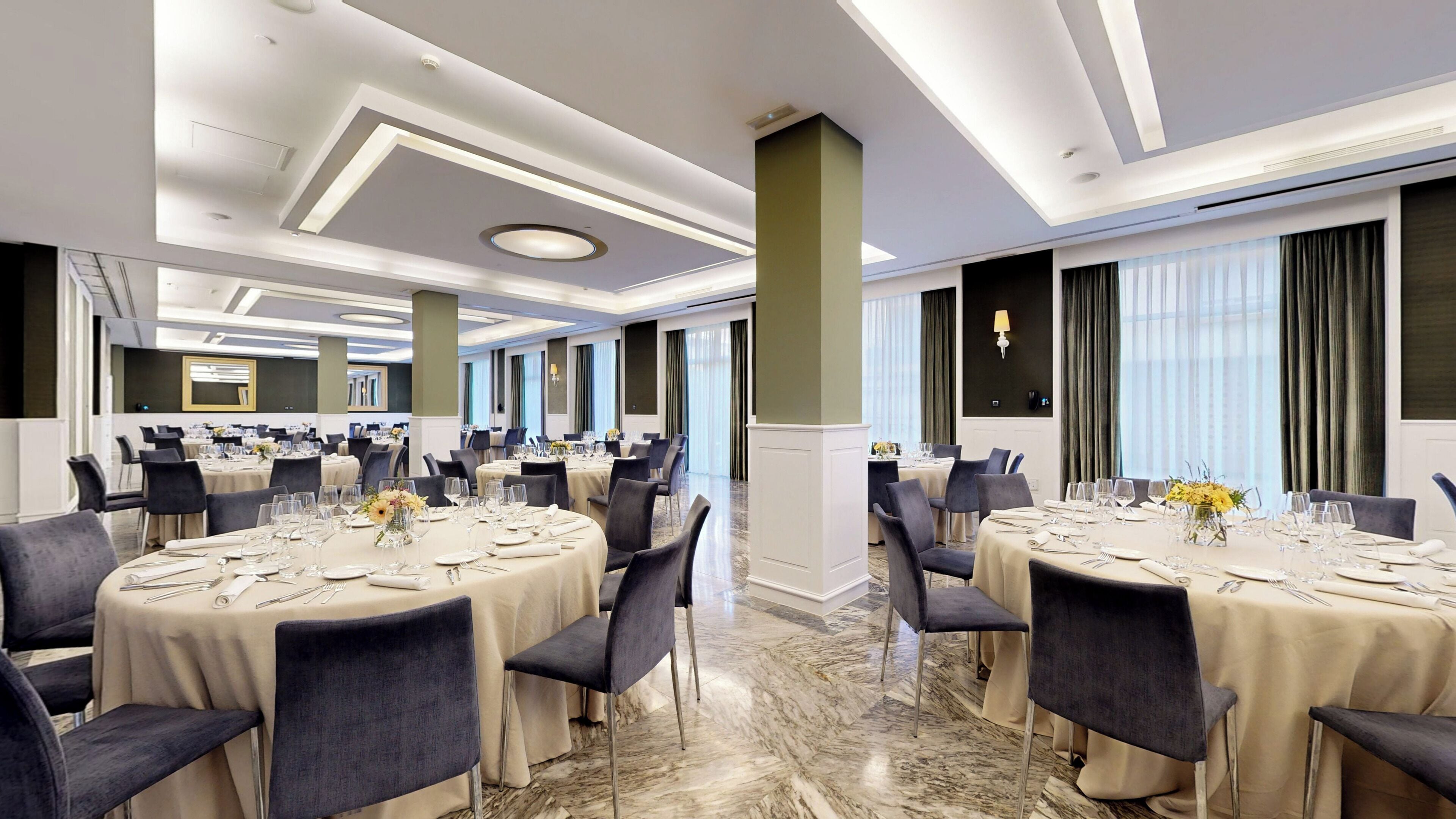 Un vaste espace de restauration d’hôtel, doté de nombreuses tables rondes dressées pour le repas, de grandes fenêtres avec des rideaux et d’un design de plafond moderne.