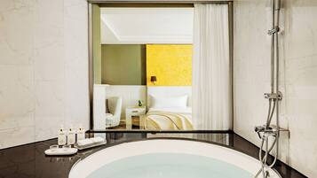 A modern bathroom with a large bathtub, a shower, and a mirror reflecting a room with a bed and a nightstand.