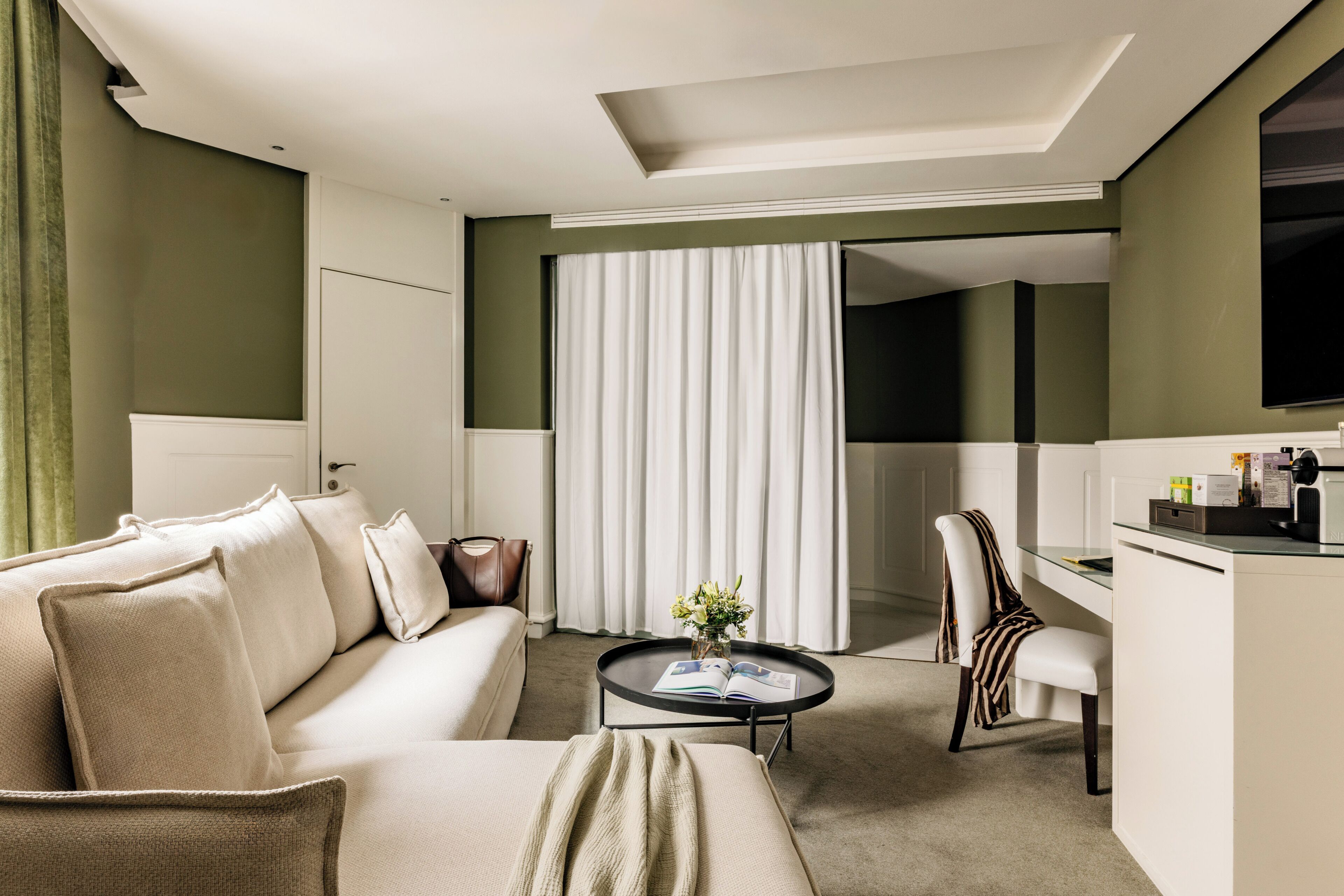 Une chambre d’hôtel moderne, équipée d’un canapé, d’une petite table basse, d’un fauteuil, d’un bureau et d’une grande fenêtre avec des rideaux.