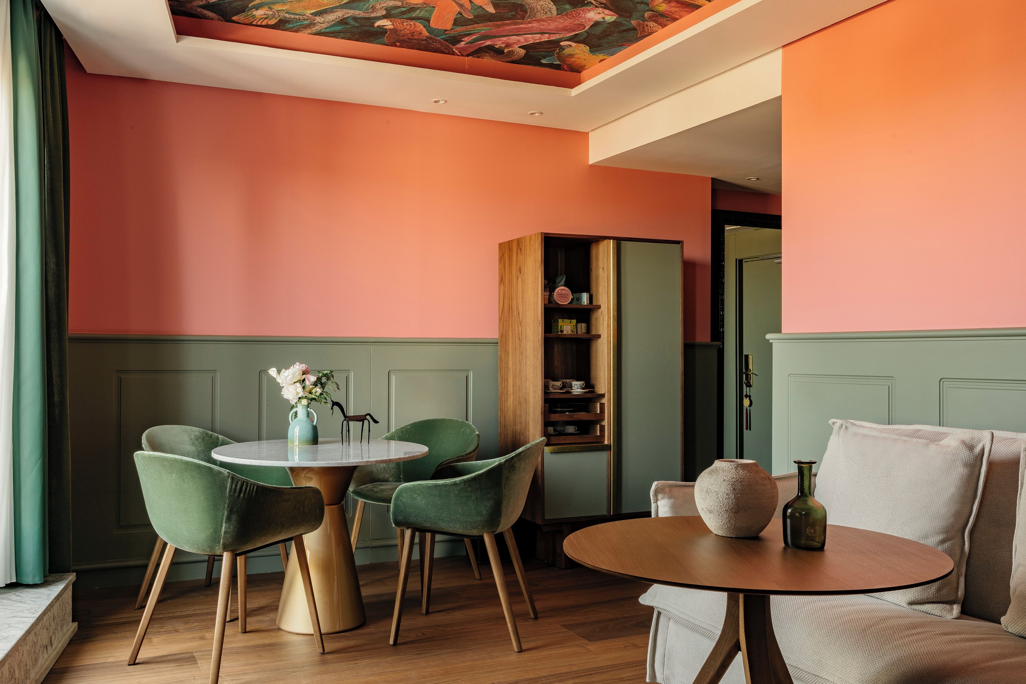 A dining area with a round table, two green chairs, a vase with flowers, and a wooden cabinet with shelves.
