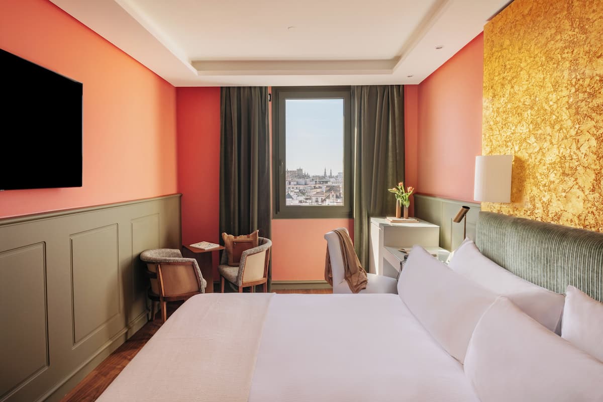 A hotel room with a large bed, a desk, a chair, and a view of a cityscape through a window.
