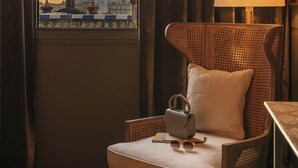 A room with a view of a cityscape through a window, a wicker chair with a cushion, a small table with a lamp, and a handbag on the chair.