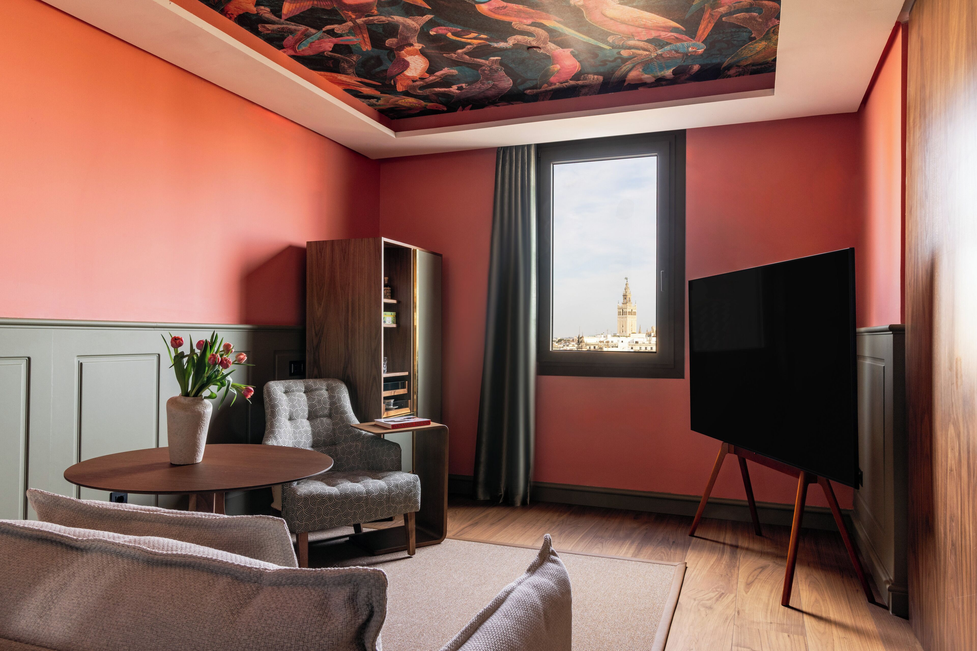A room with a large window, a flat-screen TV, a sofa, a chair, and a bookshelf.