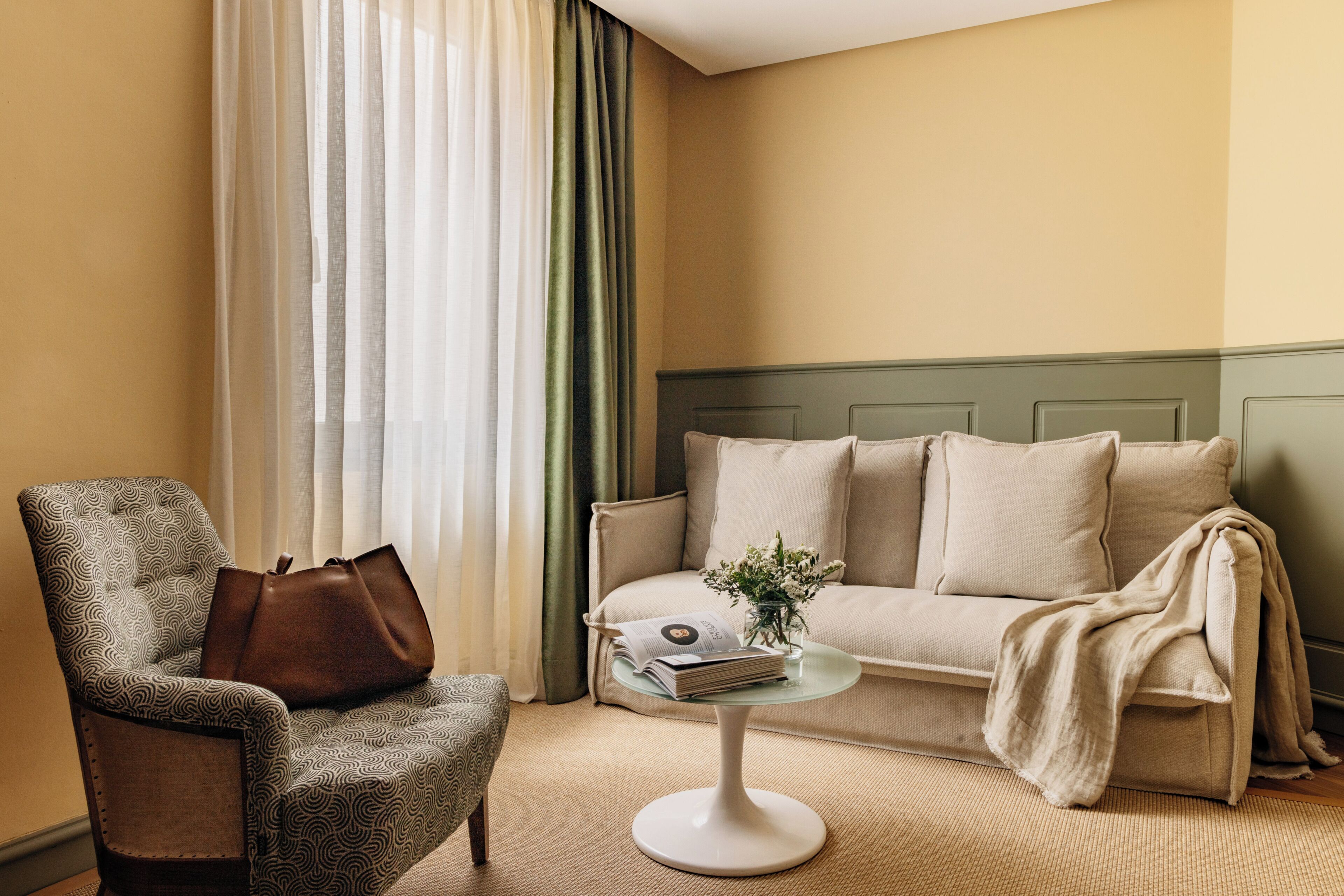 Una sala de estar con un sillón estampado, un sofá, una mesita redonda con un libro y un florero, y una ventana con cortinas translúcidas.