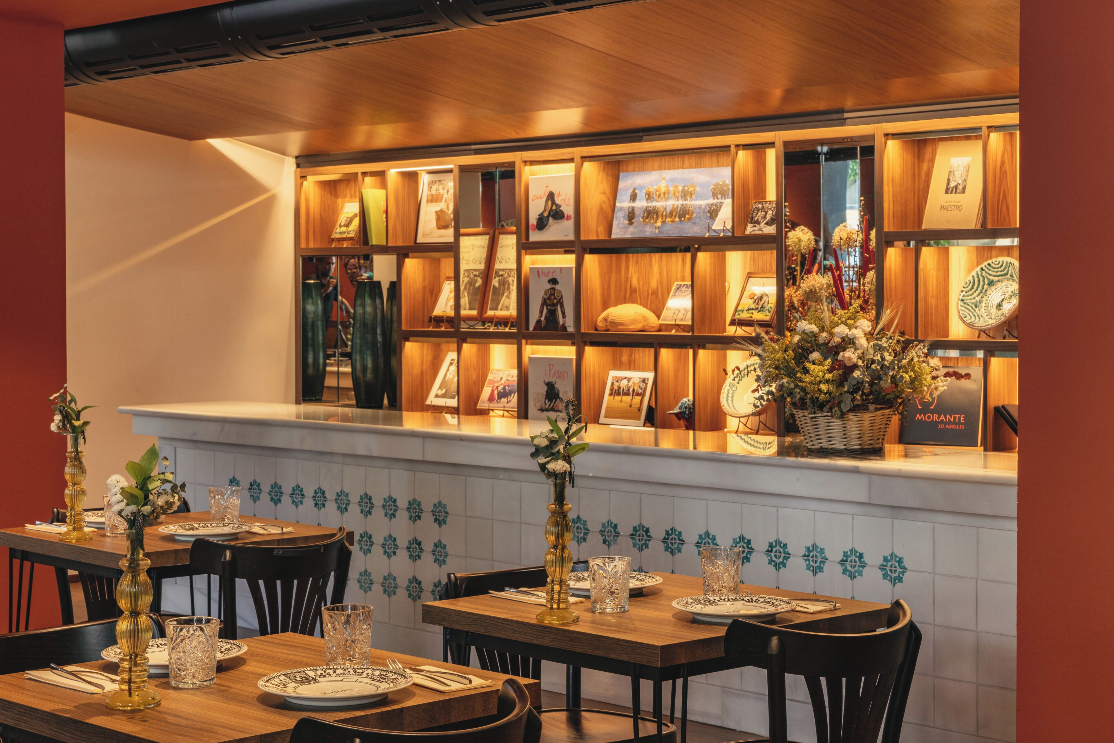 A restaurant interior with wooden tables, chairs, a tiled backsplash, and a display of framed pictures and decorative items.
