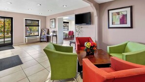 A hotel lobby with green and red chairs, a wooden table, a TV mounted on the wall, and a framed picture.