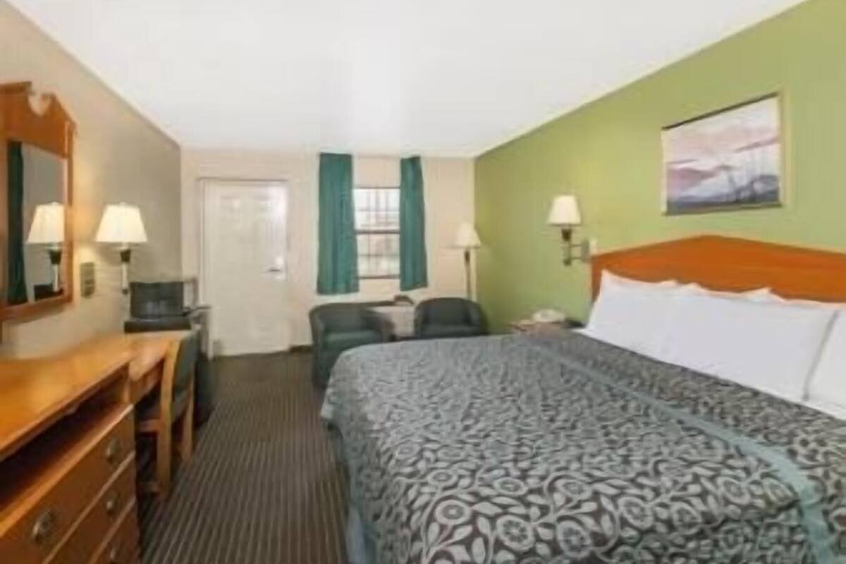 Une chambre d’hôtel comprenant un lit, un bureau, une chaise, une commode, un miroir, une porte et une fenêtre avec des rideaux.