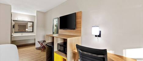 Habitación de hotel compacta con cama, escritorio y silla, televisor de pantalla plana en la pared, microondas y pequeña zona de kitchenette.