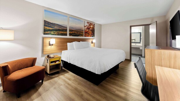 A modern hotel room with a large bed, a wooden nightstand, a brown armchair, a desk, and a flat-screen TV.
