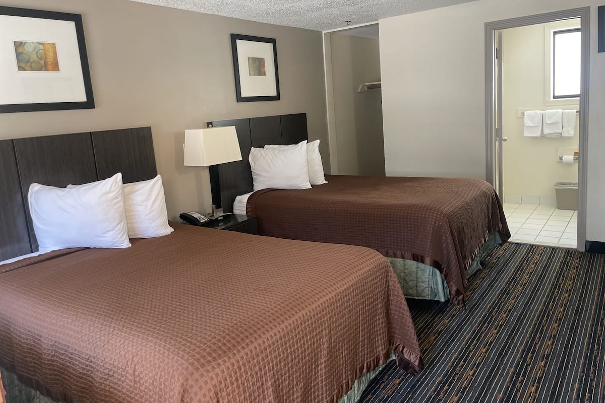A hotel room with two beds, a nightstand with a lamp, a framed picture on the wall, and a bathroom visible through an open door.
