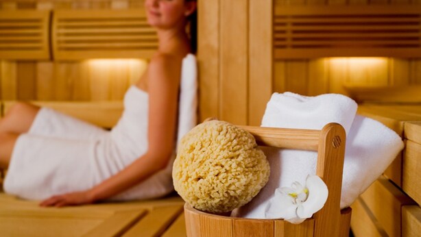 A person in a white bathrobe sitting in a wooden sauna with a towel and a round bath mat on a bench. - Romantik Hotel Bülow Residenz (Dresden)