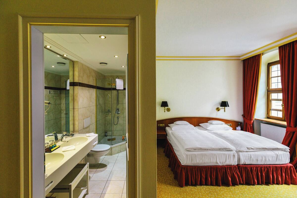 A hotel room with a bathroom, a bed with red curtains, and a window with red curtains.