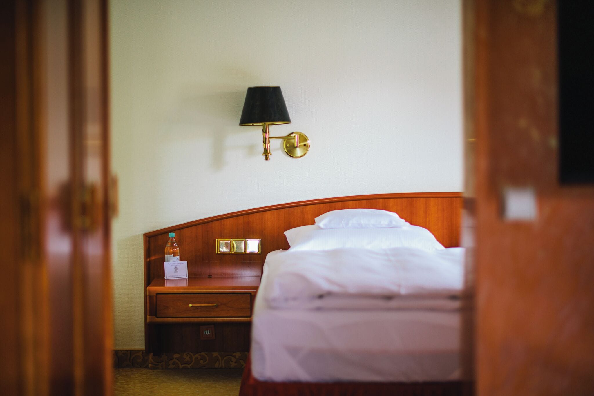 Ein Hotelzimmer mit einem hölzernen Bett, einem Nachttisch mit einer Flasche und einer Karte sowie einer Wandlampe.