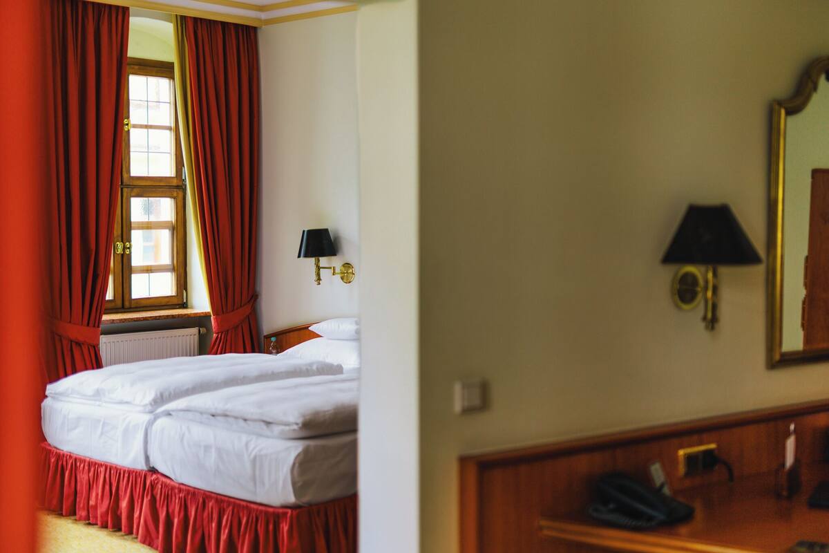 A hotel room with a bed, a window with red curtains, a lamp, and a mirror.