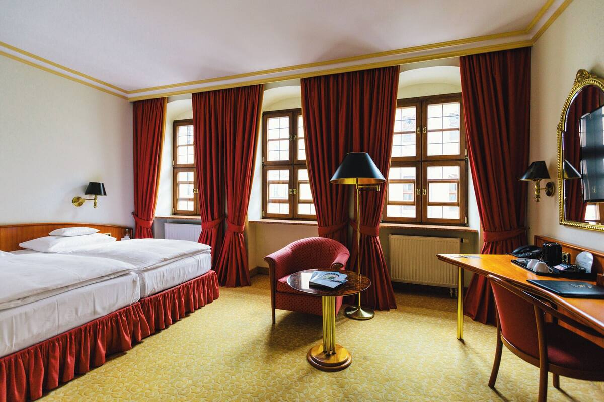 A hotel room with a bed, a desk, a chair, a lamp, and a window with curtains.