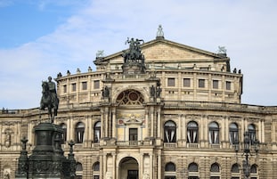 Un edificio histórico con arquitectura clásica, que presenta un frontón central, varias estatuas y una entrada destacada.