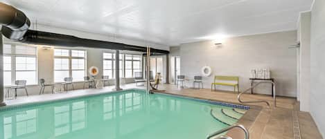 Une piscine intérieure à l’eau claire, entourée de chaises et de tables, et dotée de grandes fenêtres qui laissent entrer la lumière naturelle.