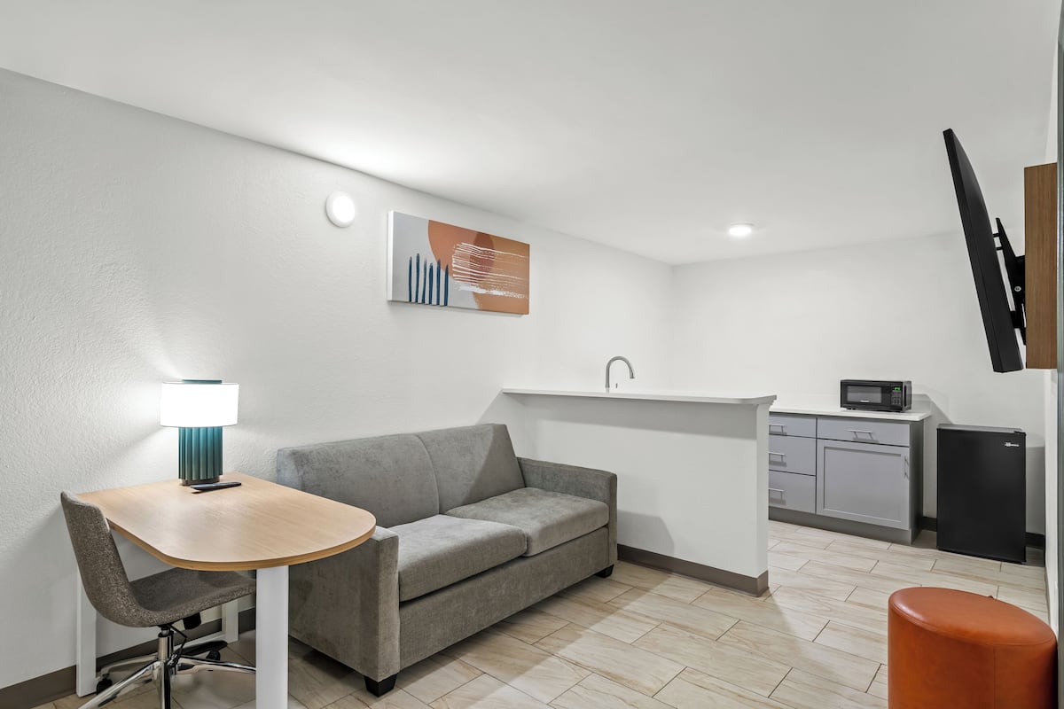 Ett kompakt bostadsutrymme med en soffa, ett litet bord med en lampa, en liten köksdel och en platt-TV som är monterad på väggen.