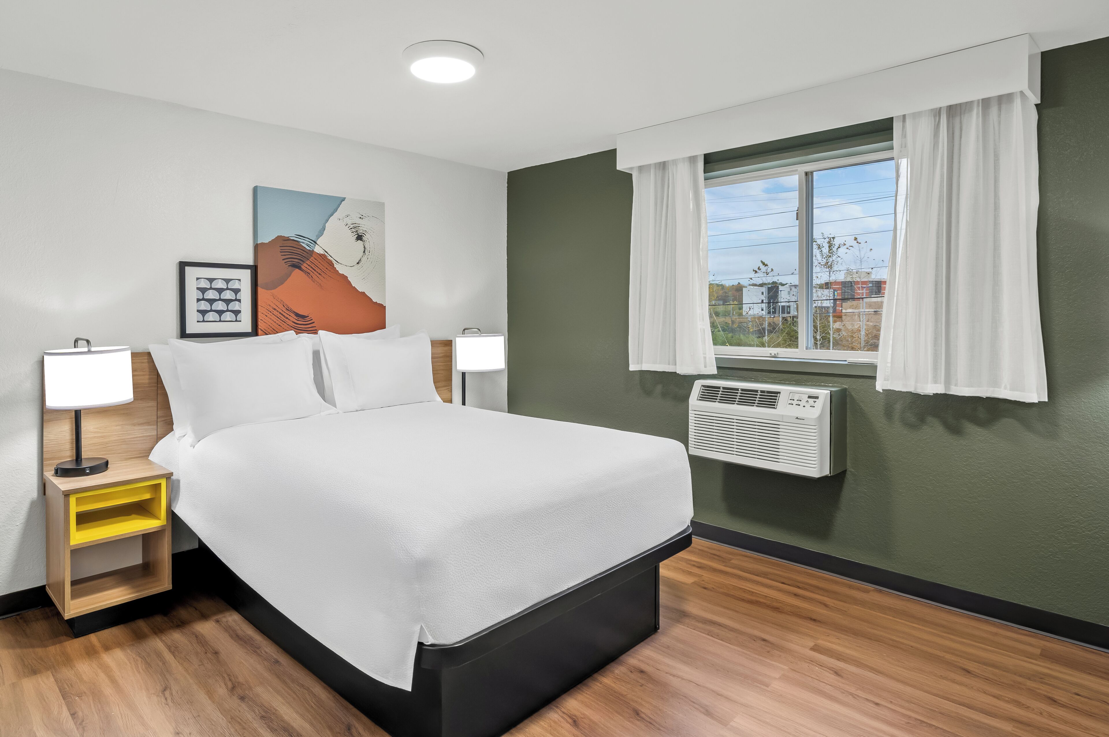 A neatly arranged hotel room with a bed, bedside tables, a window with curtains, and an air conditioning unit.