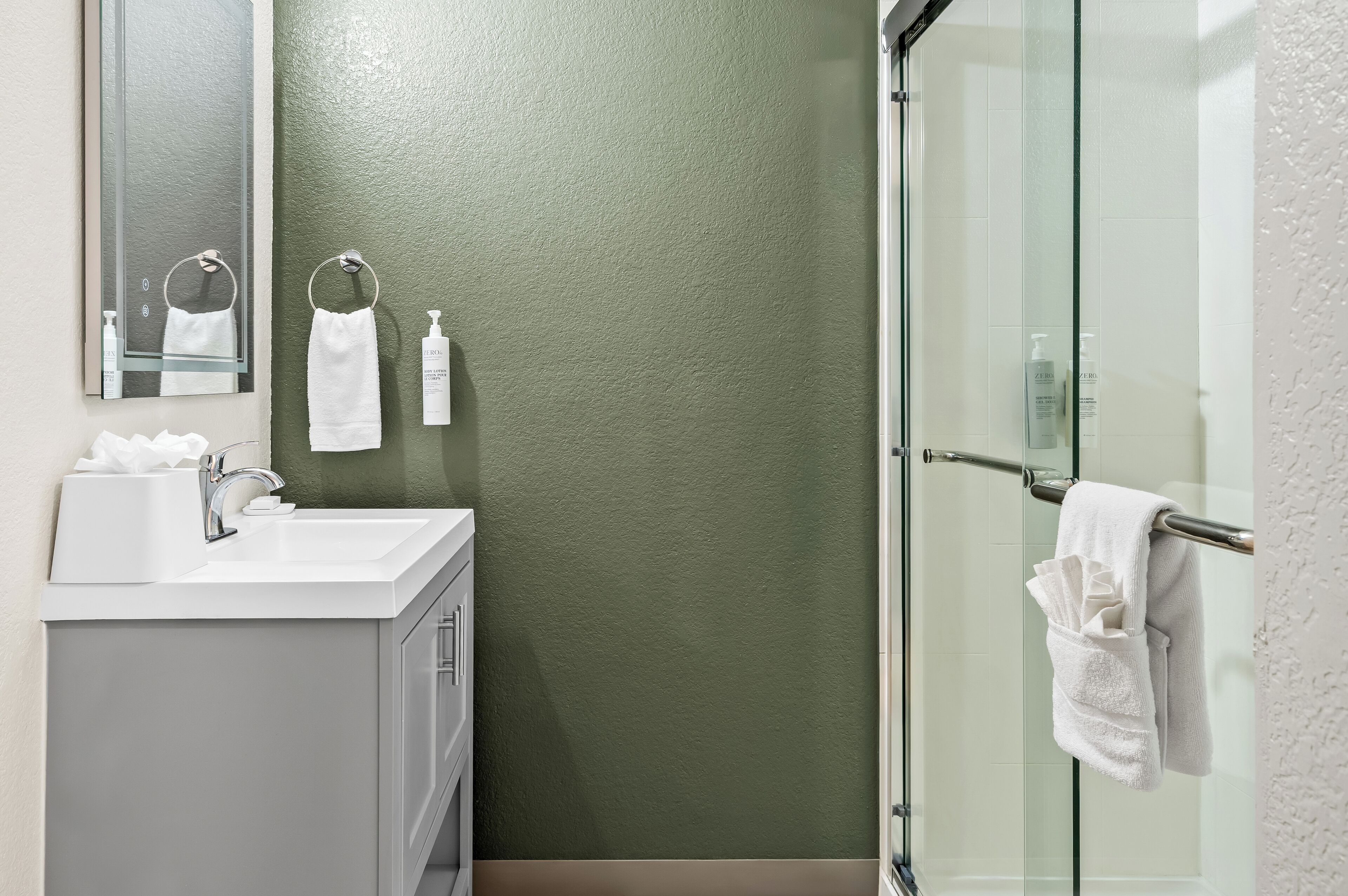 A modern bathroom with a glass shower enclosure, a white sink, and a green textured wall.