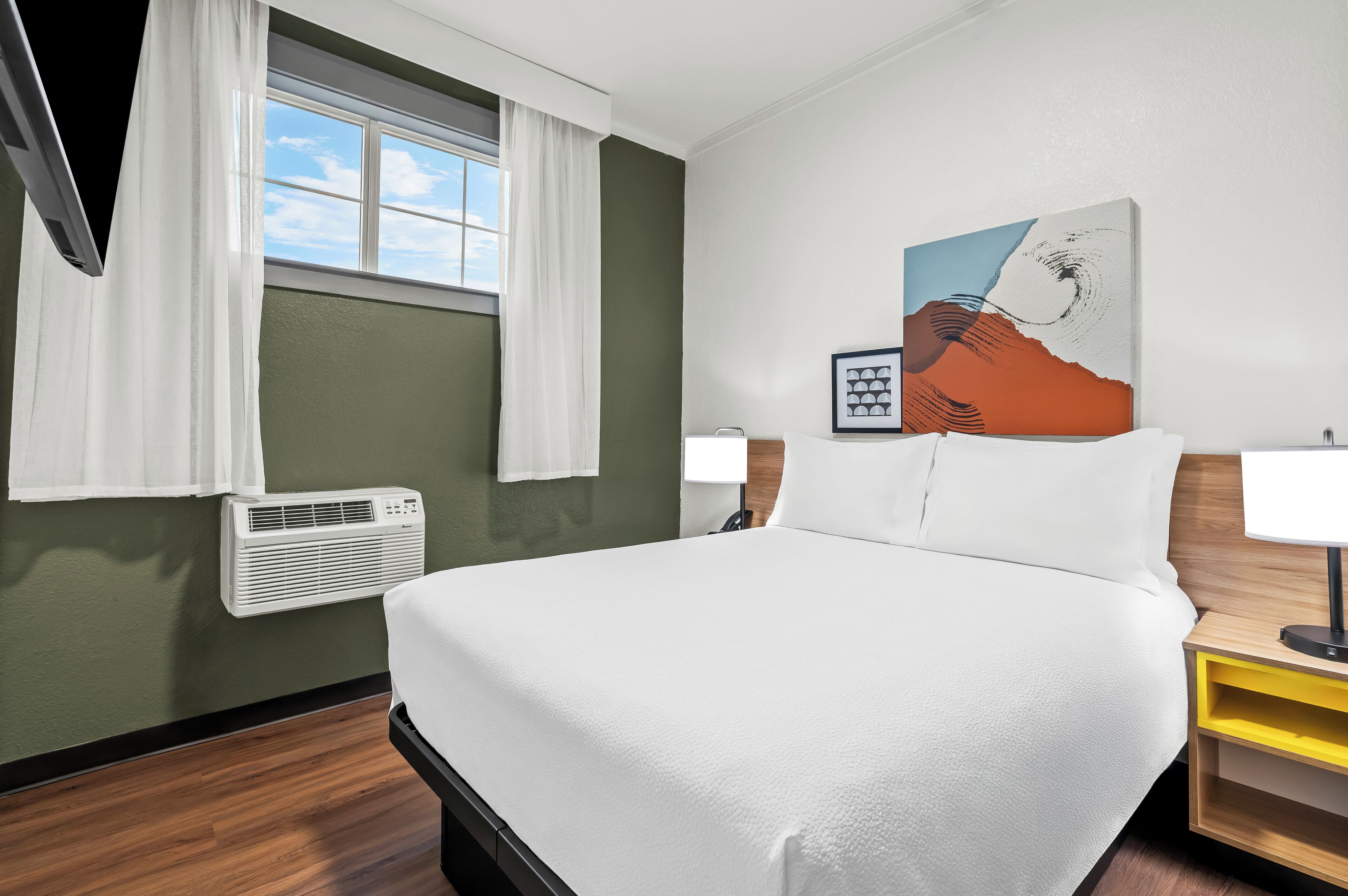 Ein Hotelzimmer mit einem großen Bett, einem Fenster mit Vorhängen, einer Klimaanlage, einem Bild an der Wand und einem Nachttisch mit Lampe.