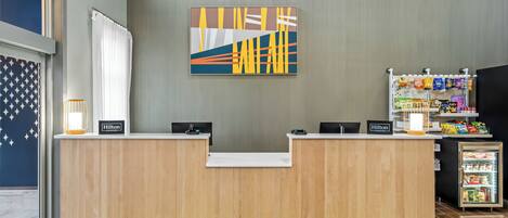 Balcão de recepção de hotel com design moderno, uma obra de arte na parede e uma máquina de venda de lanches.