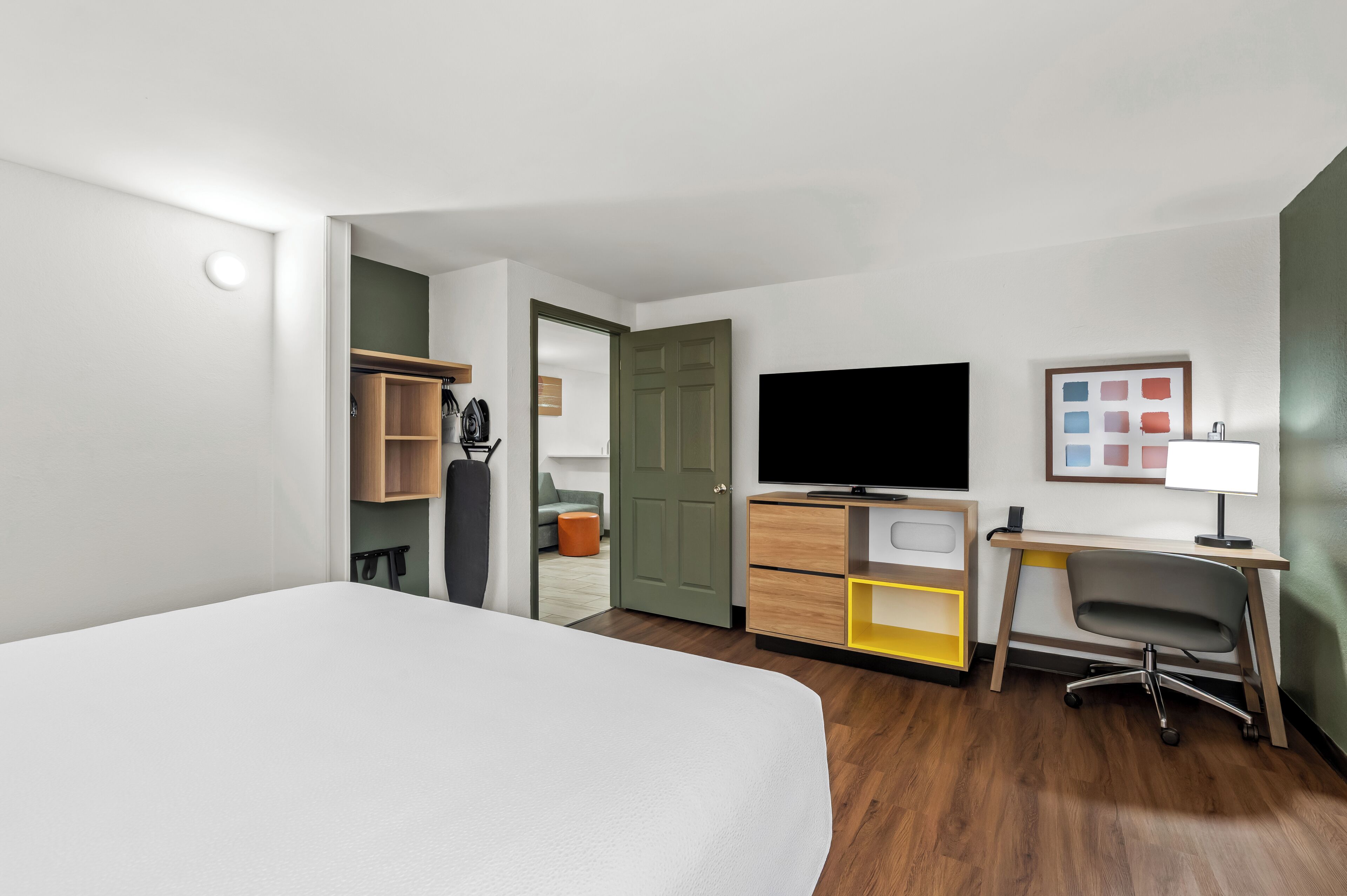 A hotel room with a bed, a desk with a chair, a television, a wardrobe, and a door leading to another room.