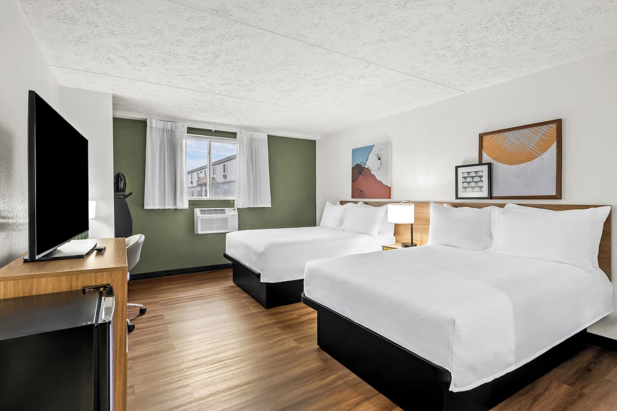 Ett hotellrum med två sängar, ett skrivbord, en platt-TV, ett fönster med gardiner och konstverk på väggarna.