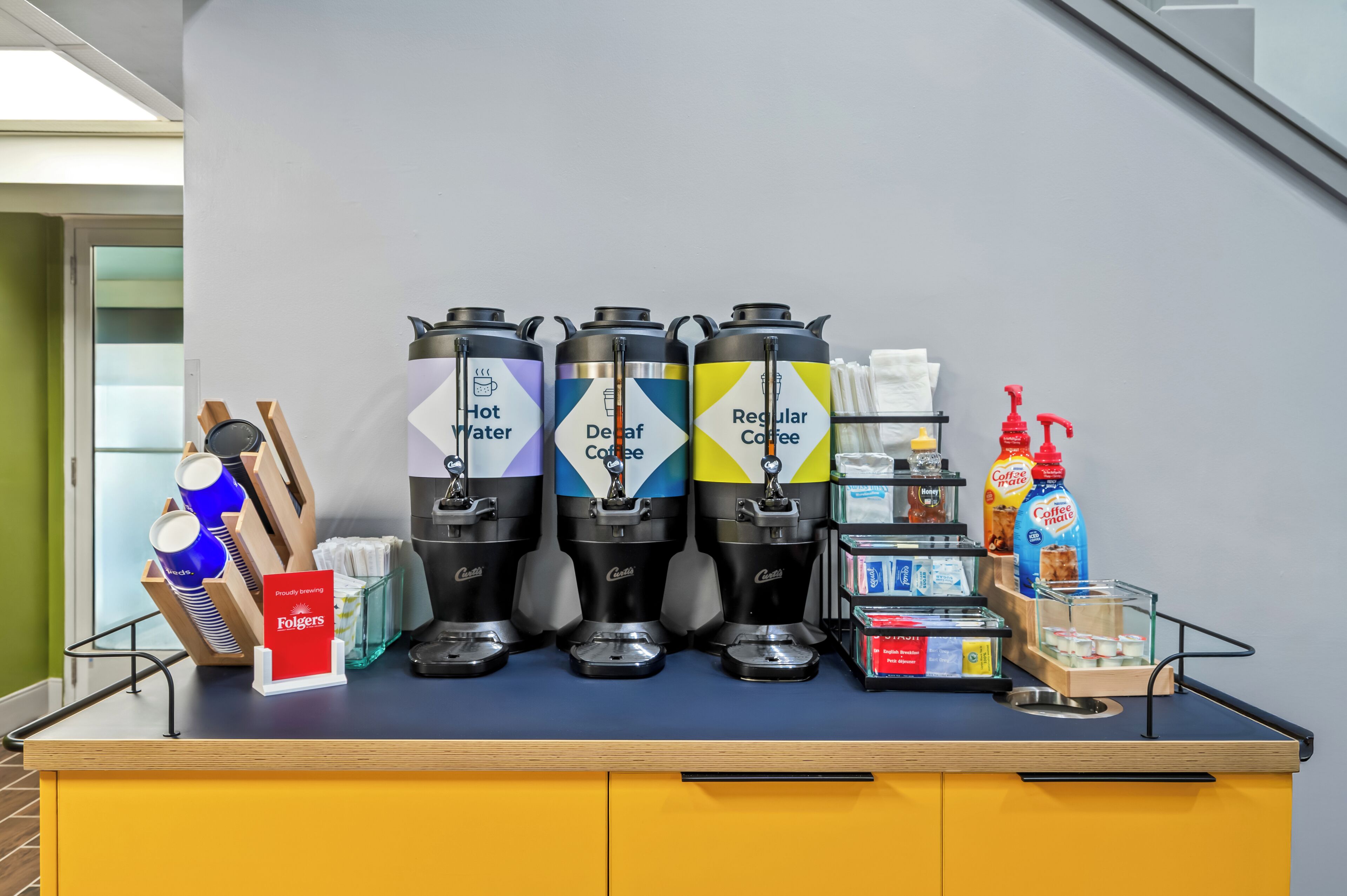 Un espace dédié au café, équipé d’un distributeur d’eau chaude, d’un distributeur de café décaféiné et d’un distributeur de café ordinaire, ainsi que d’une machine à café et d’une bouteille de crème à café.