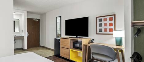 Habitación de hotel con cama, escritorio con computadora, televisor, microondas, un mueble pequeño y un baño visible a través de una puerta abierta.