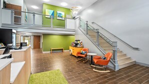 Un vestíbulo de hotel con mostrador de recepción, una pared verde, una escalera y una zona de estar con sillas anaranjadas.