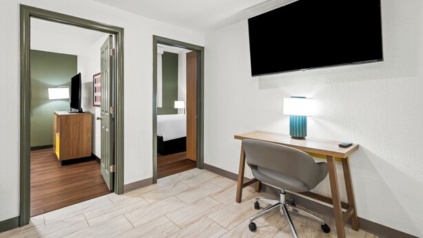 Habitación de hotel con un escritorio de madera, una silla, un televisor montado en la pared y una cama en la habitación contigua.