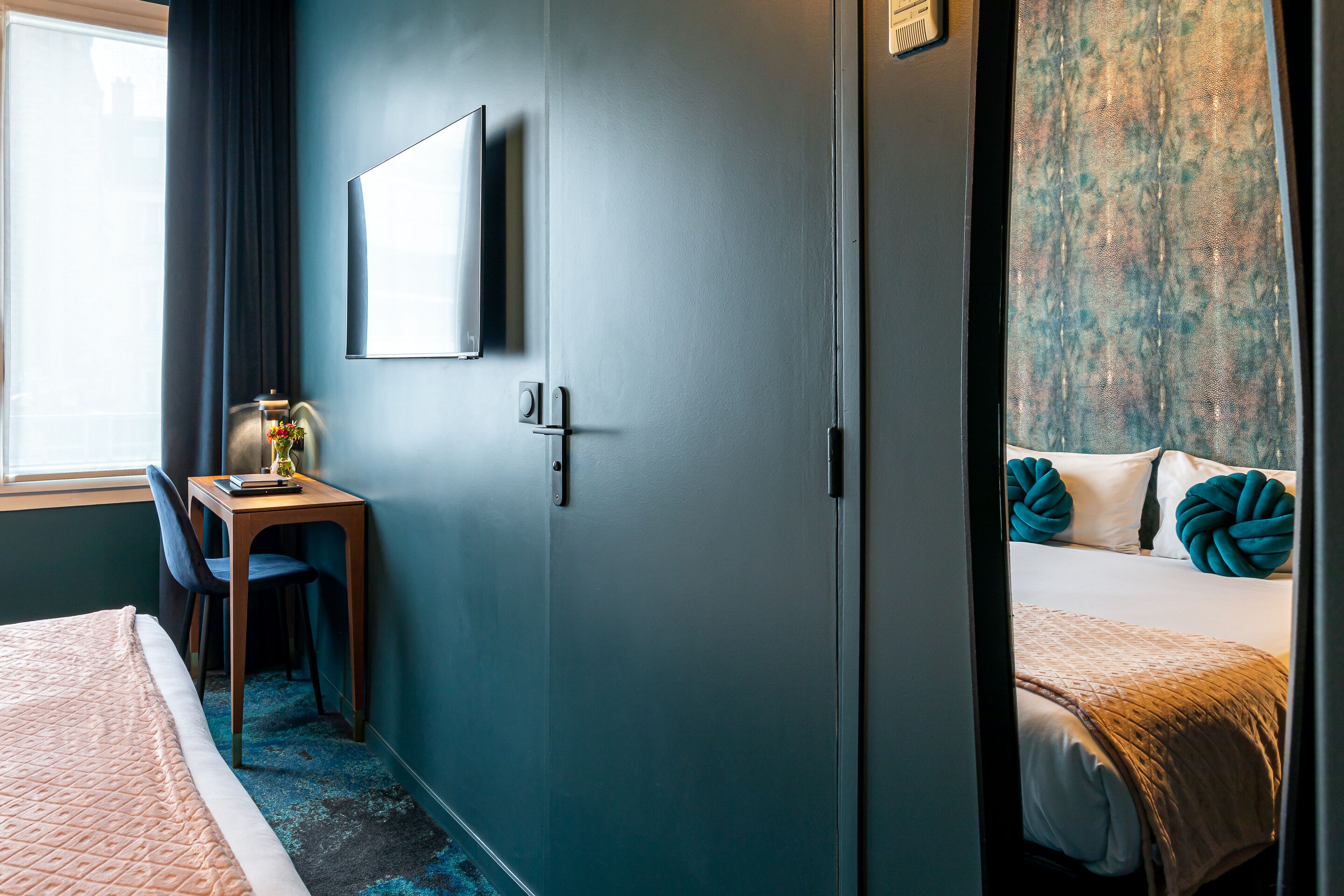 A hotel room with a bed, a desk with a chair, a TV mounted on the wall, and a window with curtains.