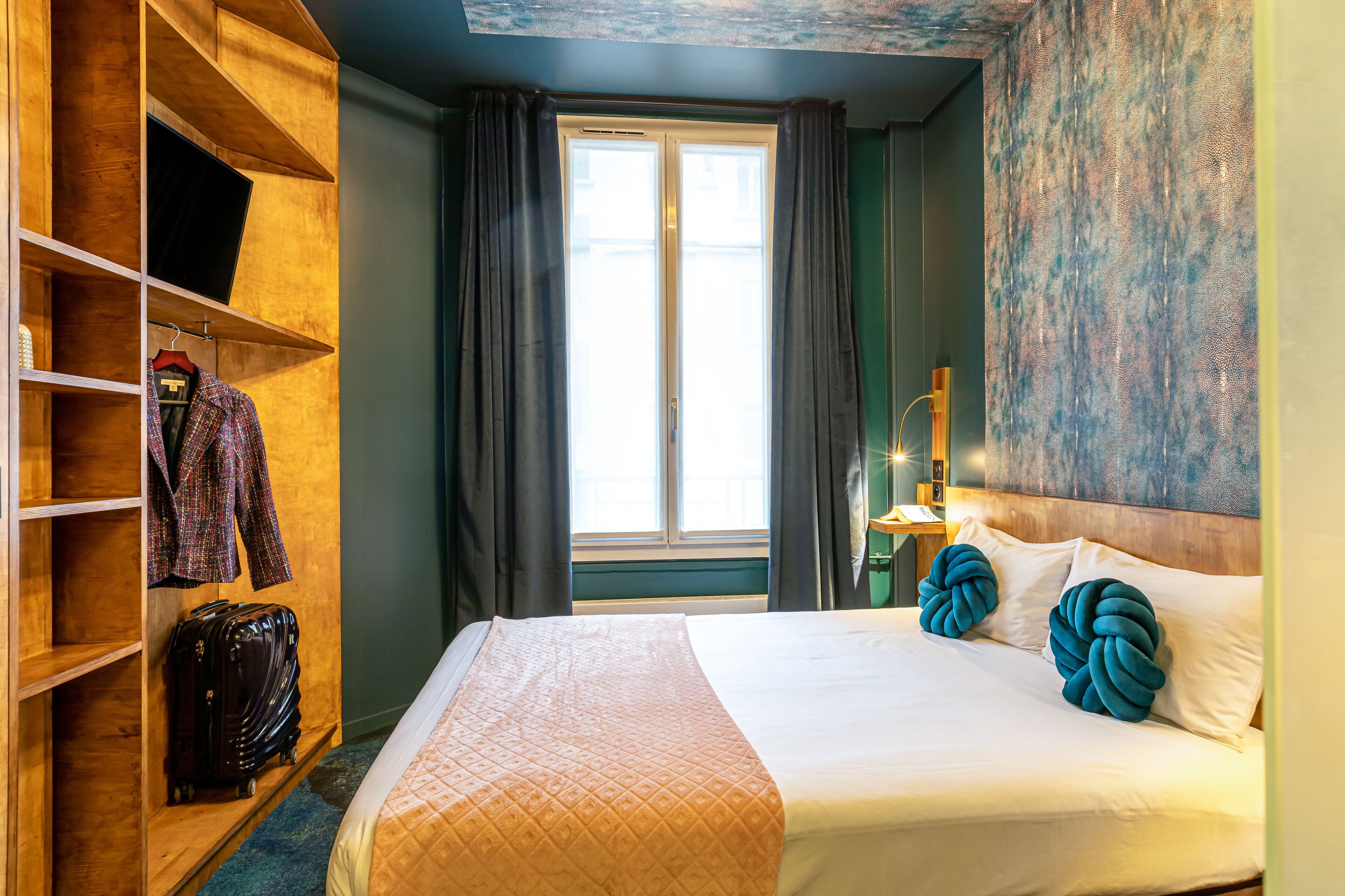 Habitación de hotel con cama, armario, maleta, ventana con cortinas y pared con diseño texturizado.